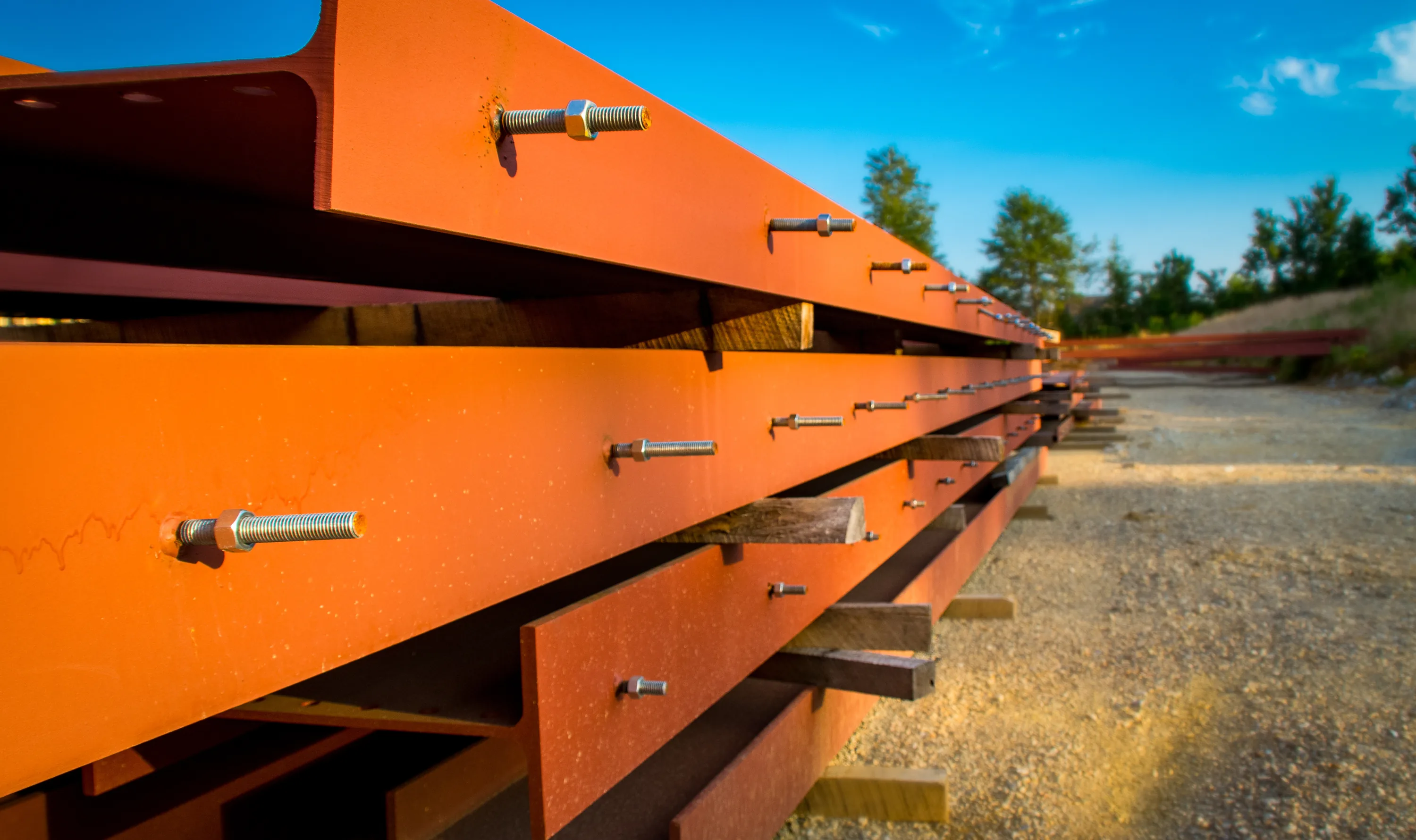 Stacked orange-primed steel I-beams with anchor bolts, lit by low evening sun
